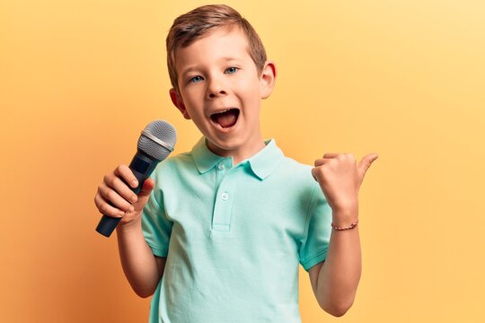 Cute blond kid singing song using microphone pointing thumb up to the side smiling happy with open mouth