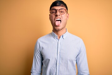 Young handsome hispanic business man wearing nerd glasses over yellow background sticking tongue out happy with funny expression. Emotion concept.