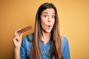 Young beautiful girl holding healthy protein bar standing over isolated yellow background scared in...