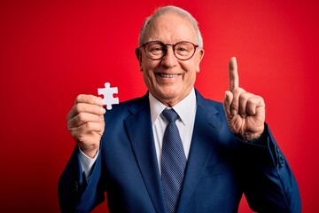 Grey haired senior business man holding puzzle piece as union and cooperation surprised with an idea or question pointing finger with happy face, number one