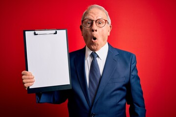 Senior grey haired business man holding clipboard over red background scared in shock with a...