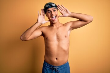 Young handsome man shirtless wearing swimsuit and swim cap over isolated yellow background Smiling cheerful playing peek a boo with hands showing face. Surprised and exited