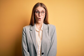 Young beautiful redhead woman wearing jacket and glasses over isolated yellow background afraid and...