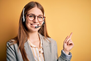 Young redhead call center agent woman overworked wearing glasses using headset with a big smile on face, pointing with hand and finger to the side looking at the camera.