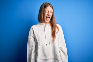 Young beautiful redhead sporty woman wearing sweatshirt over isolated blue background winking looking at the camera with sexy expression, cheerful and happy face.