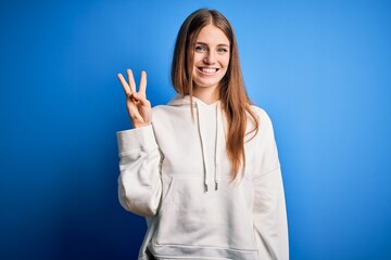 Young beautiful redhead sporty woman wearing sweatshirt over isolated blue background showing and pointing up with fingers number three while smiling confident and happy.