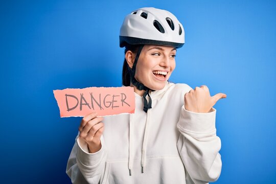 Young beautiful redhead cyclist woman wearing bike helmet holding danger paper message pointing and showing with thumb up to the side with happy face smiling