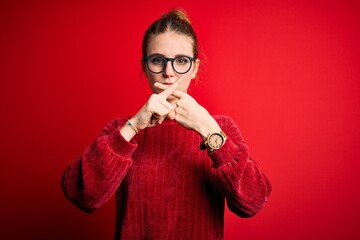 Young beautiful redhead woman wearing casual sweater over isolated red background Rejection expression crossing fingers doing negative sign
