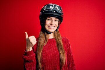Young beautiful redhead motocyclist woman wearing moto helmet over red background doing happy thumbs up gesture with hand. Approving expression looking at the camera showing success.