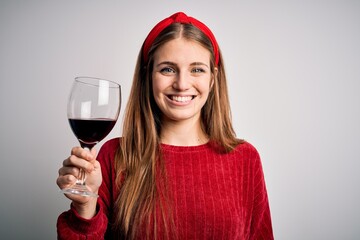 Young beautiful redhead woman drinking glass of red wine over isolated white background with a...