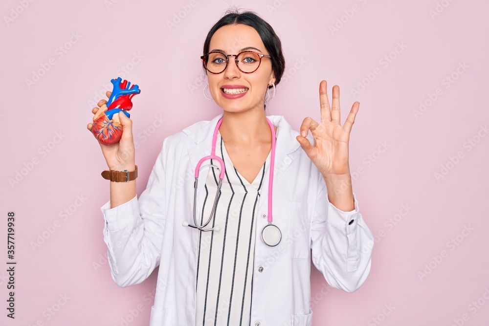 Wall mural Young beautiful cardiologist woman with blue eyes wearing stethoscope holding heart doing ok sign with fingers, excellent symbol