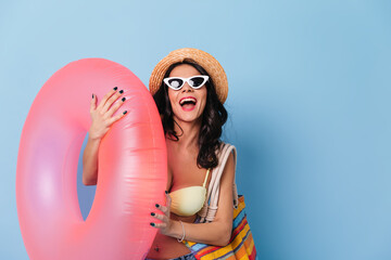 Excited lady in sunglasses holding swimming circle. Studio shot of laughing girl in straw hat isolated on blue background.