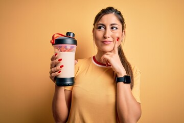 Young beautiful brunette woman holding gym bottle shaker with protein beverage for diet serious...