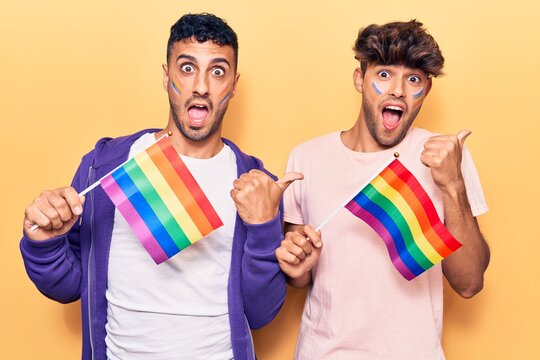 Young gay couple holding rainbow lgbtq flags pointing thumb up to the side smiling happy with open mouth