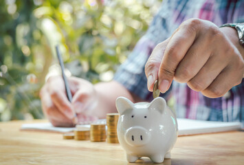 Young businessman writing receipts Expenditure into notebooks and putting coins in piggy banks to...
