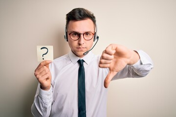 Young call center operator man wearing headset holding paper note with question mark with angry face, negative sign showing dislike with thumbs down, rejection concept