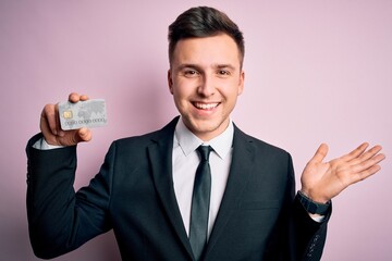 Young handsome caucasian business man holding finance credit card over pink background very happy and excited, winner expression celebrating victory screaming with big smile and raised hands