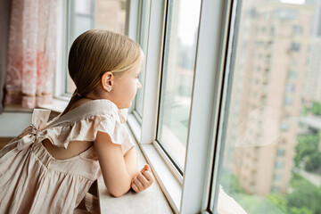 Stylish little girl with blonde hair sitting at home near window during coronavirus covid-19 self isolation
