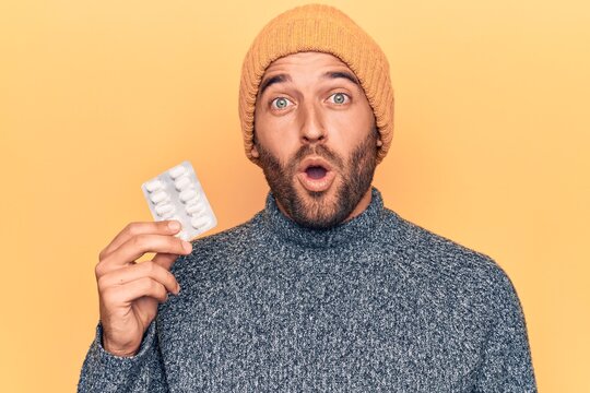 Young handsome blond man with beard holding medicine pills over yellow background scared and amazed with open mouth for surprise, disbelief face