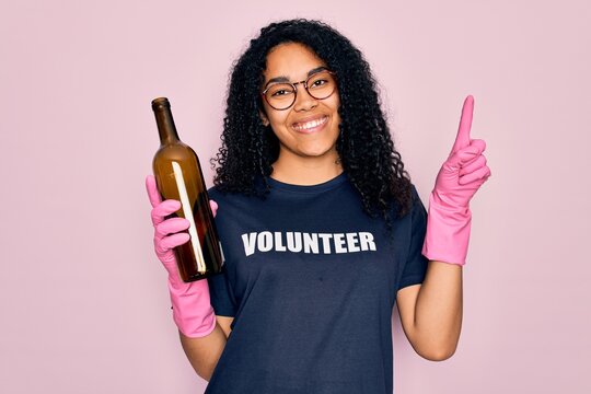African american curly woman wearing volunteer t-shirt doing volunteering recycling glass bottle surprised with an idea or question pointing finger with happy face, number one