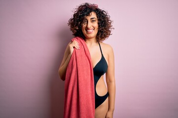 Young beautiful curly arab woman on vacation wearing swimsuit holding beach towel with a happy face standing and smiling with a confident smile showing teeth