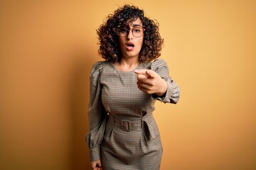 Beautiful arab business woman wearing dress and glasses standing over yellow background pointing displeased and frustrated to the camera, angry and furious with you