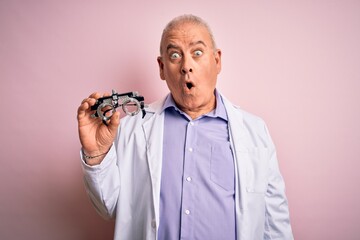 Middle age hoary optical man wearing coat holding optometry glasses over pink background scared in shock with a surprise face, afraid and excited with fear expression