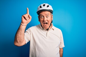 Middle age hoary cyclist man wearing bike security helmet over isolated blue background pointing finger up with successful idea. Exited and happy. Number one.
