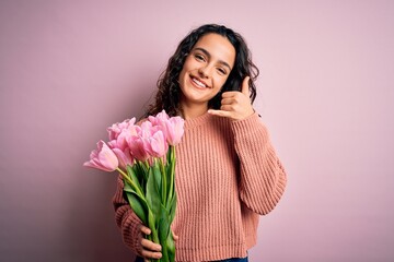 Young beautiful romantic woman with curly hair holding bouquet of pink tulips smiling doing phone...