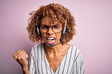 African american curly call center agent woman working using headset over pink background angry and mad raising fist frustrated and furious while shouting with anger. Rage and aggressive concept.