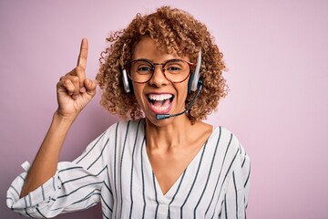 African american curly call center agent woman working using headset over pink background pointing finger up with successful idea. Exited and happy. Number one.