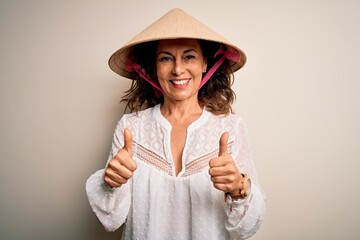 Middle age brunette woman wearing asian traditional conical hat over white background success sign doing positive gesture with hand, thumbs up smiling and happy. Cheerful expression and winner