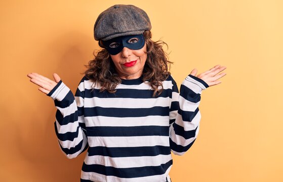 Middle age brunette burglar woman wearing mask and cap standing over yellow background clueless and confused with open arms, no idea and doubtful face.
