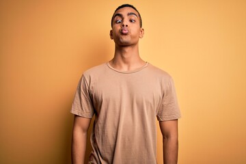 Young handsome african american man wearing casual t-shirt standing over yellow background making fish face with lips, crazy and comical gesture. Funny expression.