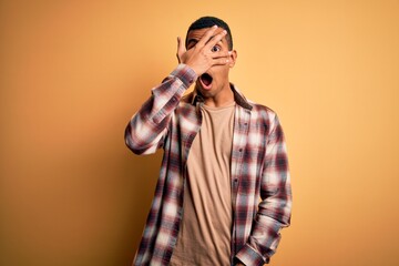 Young handsome african american man wearing casual shirt standing over yellow background peeking in shock covering face and eyes with hand, looking through fingers with embarrassed expression.