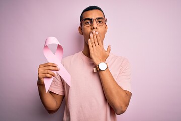 Young handsome african american man hoding pink cancer ribbon over isolated background cover mouth with hand shocked with shame for mistake, expression of fear, scared in silence, secret concept