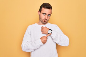 Young handsome man with blue eyes wearing casual sweater standing over yellow background In hurry...