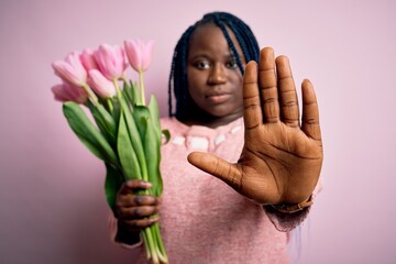 Young african american plus size woman with braids holding bouquet of pink tulips flower doing stop sing with palm of the hand. Warning expression with negative and serious gesture on the face.