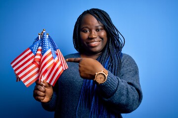 Young patriotic african american plus size woman with braids holding united states flags very happy pointing with hand and finger