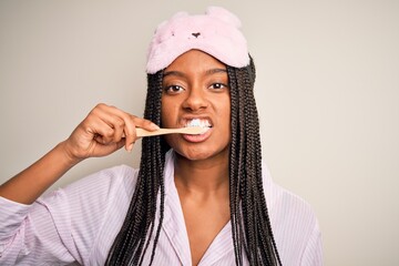 Young beautiful african american woman wearing pajama and sleep mask brushing her teeth using tooth brush and oral paste, cleaning teeth and tongue as healthy health care morning routine