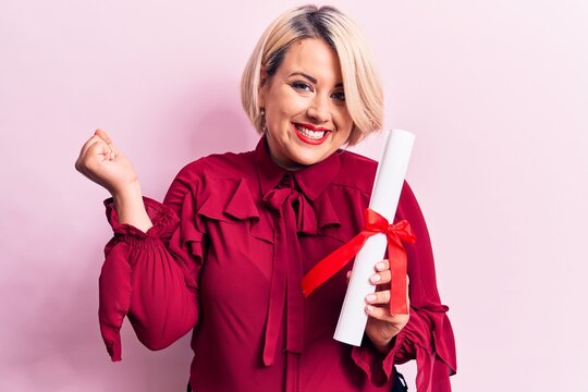 Beautiful blonde plus size woman holding graduated degree diploma over pink background screaming proud, celebrating victory and success very excited with raised arm