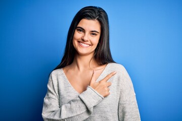 Fototapeta premium Young beautiful brunette woman wearing casual sweater standing over blue background cheerful with a smile of face pointing with hand and finger up to the side with happy and natural expression on face