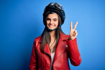 Young beautiful brunette motorcycliste woman wearing motorcycle helmet and jacket showing and pointing up with fingers number two while smiling confident and happy.