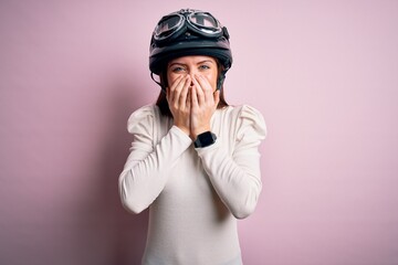 Young beautiful motorcyclist woman with blue eyes wearing moto helmet over pink background laughing...