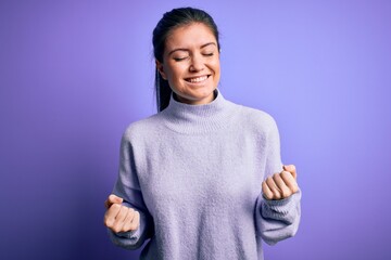 Young beautiful woman with blue eyes wearing casual turtleneck sweater over pink background very happy and excited doing winner gesture with arms raised, smiling and screaming for success. Celebration