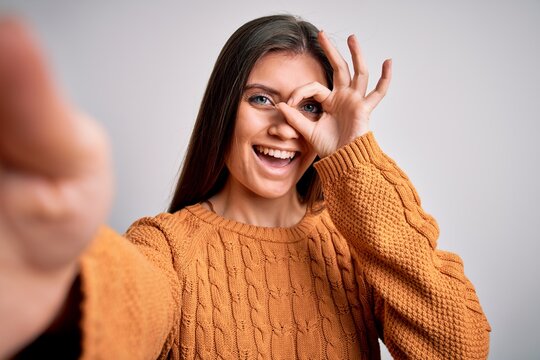 Young beautiful woman with blue eyes wearing casual sweater making selfie by camera with happy face smiling doing ok sign with hand on eye looking through fingers