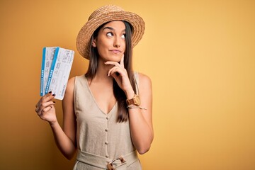Young beautiful brunette tourist woman on vacation wearing hat holding boarding pass serious face...