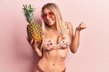 Young beautiful blonde woman wearing bikini and sunglasses holding pineapple screaming proud, celebrating victory and success very excited with raised arm