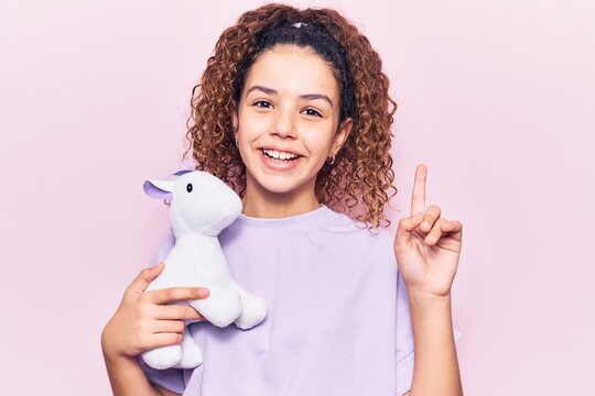 Beautiful kid girl with curly hair holding animal doll toy surprised with an idea or question pointing finger with happy face, number one
