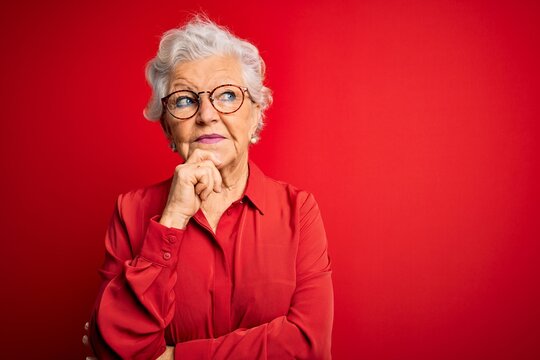 Senior beautiful grey-haired woman wearing casual shirt and glasses over red background with hand on chin thinking about question, pensive expression. Smiling with thoughtful face. Doubt concept.